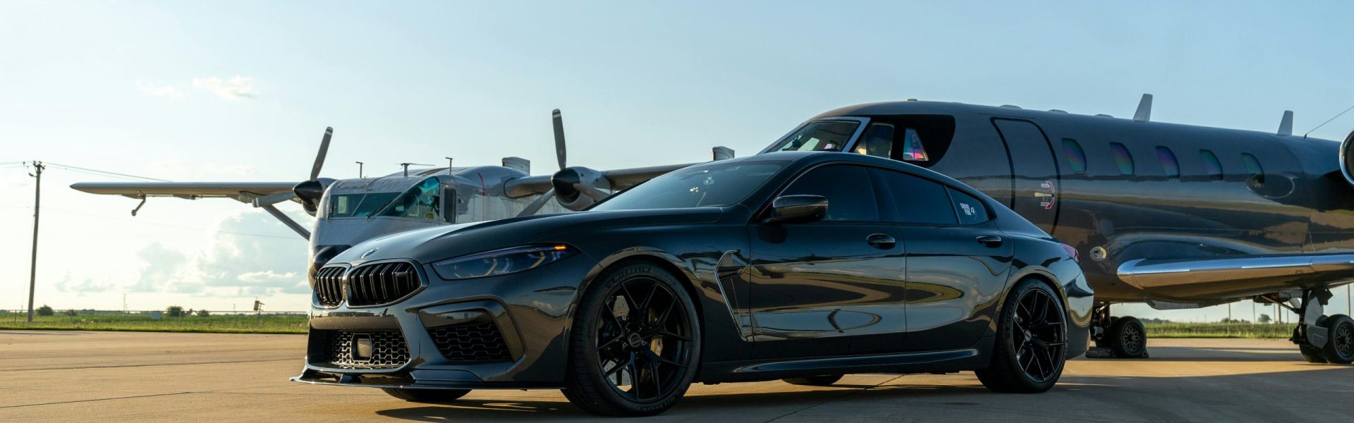 Luxury black car parked alongside a sleek private jet on a sunny airport tarmac.
